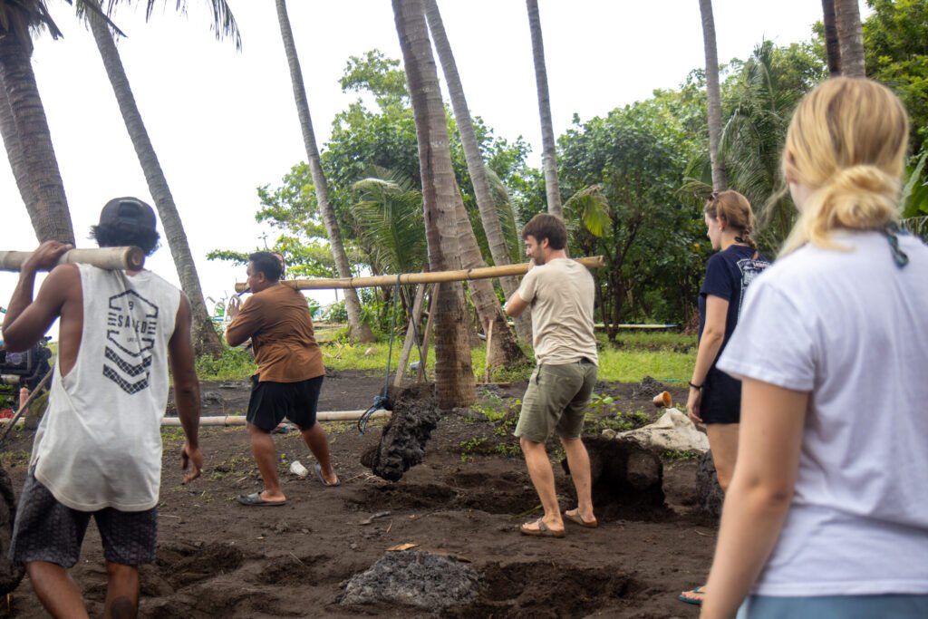 Voluntários e membros da equipe local carregando postes de bambu em uma praia durante trabalho de restauração costeira